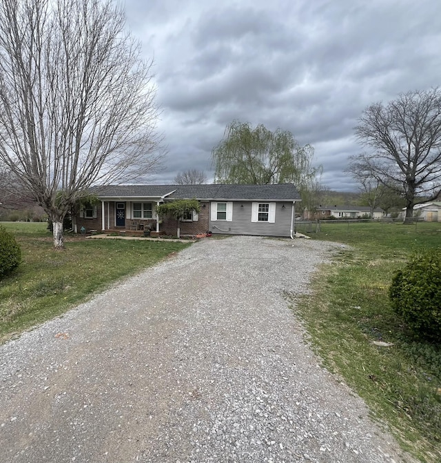 ranch-style home with gravel driveway, a front yard, brick siding, covered porch, and a shingled roof