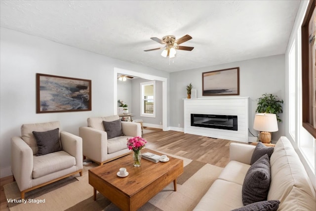 living area with wood finished floors, a glass covered fireplace, a textured ceiling, baseboards, and ceiling fan