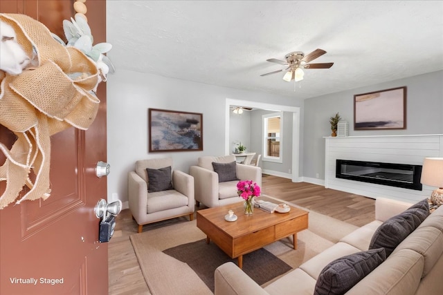 living area featuring baseboards, light wood-type flooring, a glass covered fireplace, and a ceiling fan