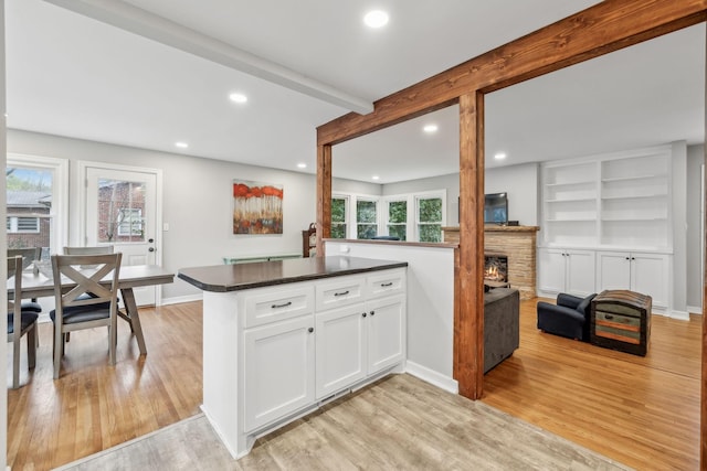kitchen with beamed ceiling, dark countertops, a lit fireplace, white cabinetry, and light wood-style floors