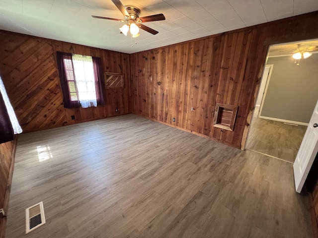 empty room with baseboards, visible vents, a ceiling fan, wood finished floors, and wooden walls