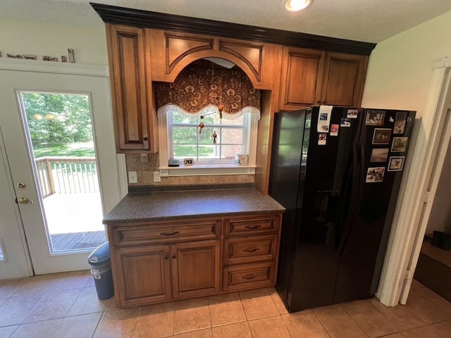 kitchen with tasteful backsplash, black fridge, brown cabinets, light tile patterned floors, and dark countertops
