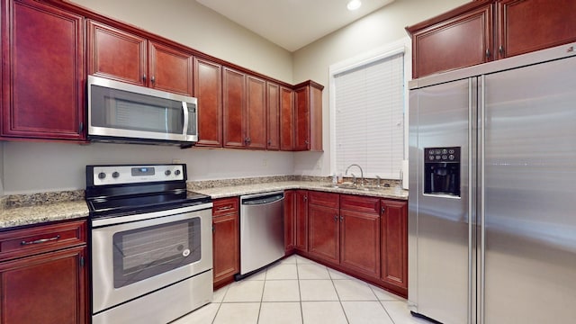 kitchen with light tile patterned floors, stainless steel appliances, a sink, reddish brown cabinets, and light stone countertops