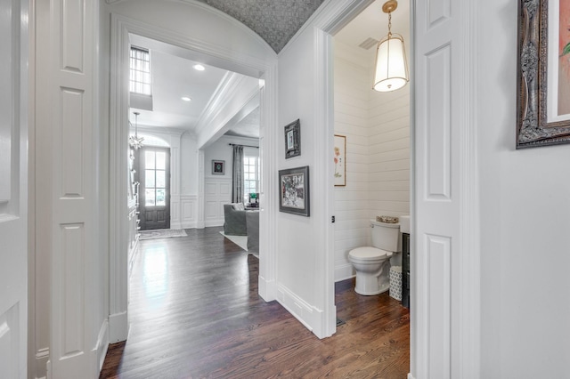corridor with a decorative wall, crown molding, an inviting chandelier, baseboards, and dark wood-style flooring