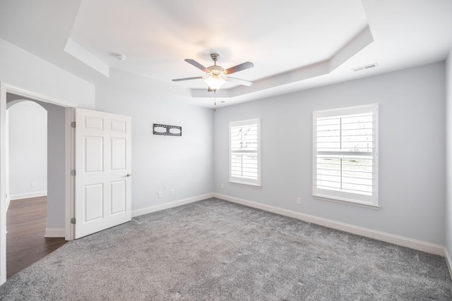carpeted spare room with visible vents, baseboards, ceiling fan, a raised ceiling, and arched walkways