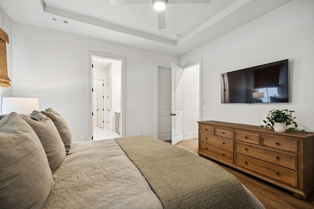 bedroom featuring a tray ceiling, connected bathroom, visible vents, a ceiling fan, and wood finished floors