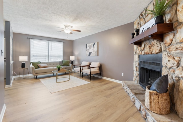 living area featuring light wood-style flooring, baseboards, a textured ceiling, a ceiling fan, and a stone fireplace