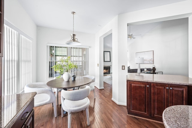 dining space with a warm lit fireplace, dark wood-style flooring, baseboards, and ceiling fan