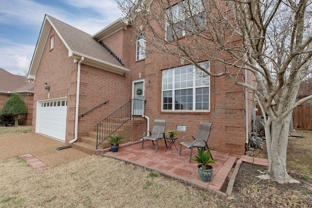 rear view of property with driveway, a patio area, roof with shingles, an attached garage, and brick siding