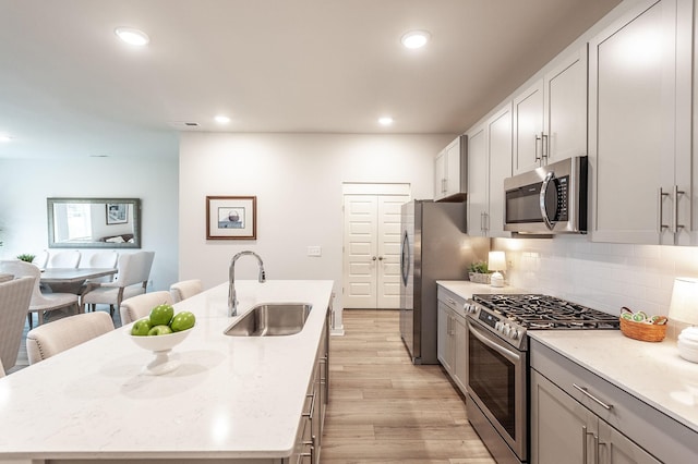 kitchen featuring an island with sink, a sink, light wood-type flooring, stainless steel appliances, and decorative backsplash