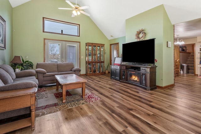 living room with high vaulted ceiling, baseboards, wood finished floors, ceiling fan with notable chandelier, and a glass covered fireplace
