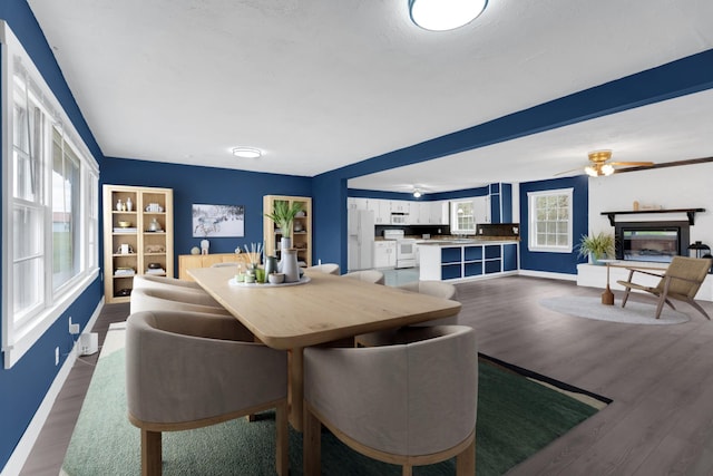 dining area featuring baseboards, a ceiling fan, a glass covered fireplace, and wood finished floors