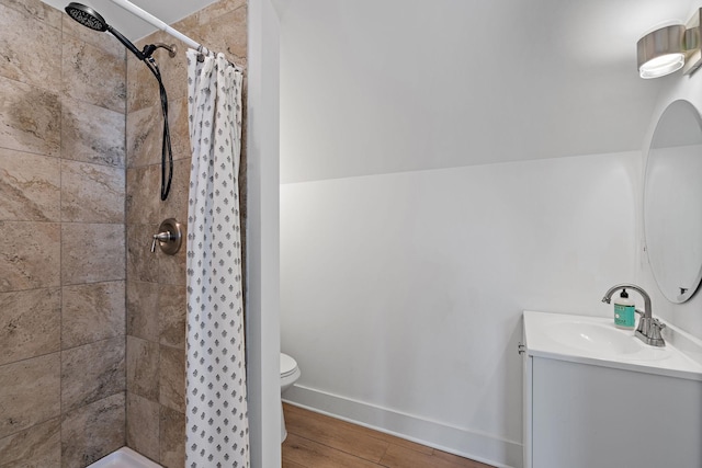 bathroom with tiled shower, vanity, toilet, wood finished floors, and baseboards