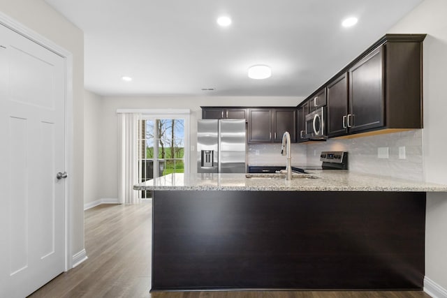 kitchen with a peninsula, dark brown cabinets, appliances with stainless steel finishes, light stone counters, and decorative backsplash