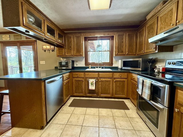 kitchen featuring a peninsula, dark countertops, under cabinet range hood, stainless steel appliances, and a sink