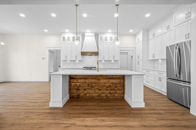 kitchen with freestanding refrigerator, dark wood finished floors, custom exhaust hood, a kitchen island with sink, and light countertops