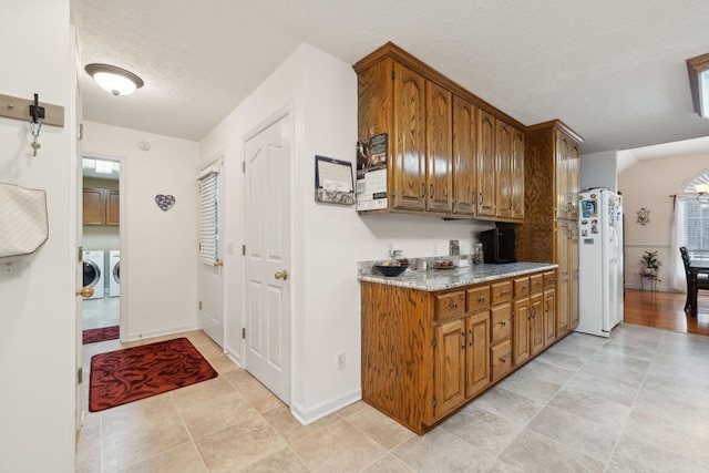 kitchen with brown cabinetry, a textured ceiling, washing machine and clothes dryer, and white fridge with ice dispenser