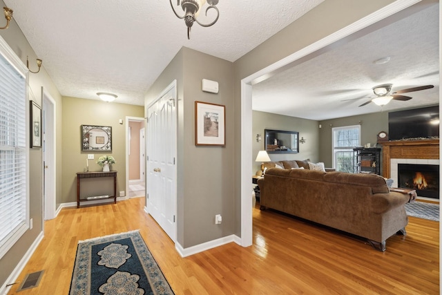 living area featuring a warm lit fireplace, a textured ceiling, visible vents, ceiling fan, and light wood finished floors