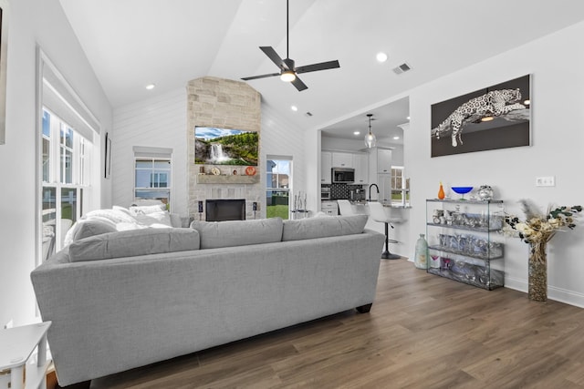 living room featuring high vaulted ceiling, ceiling fan, dark wood finished floors, a stone fireplace, and visible vents