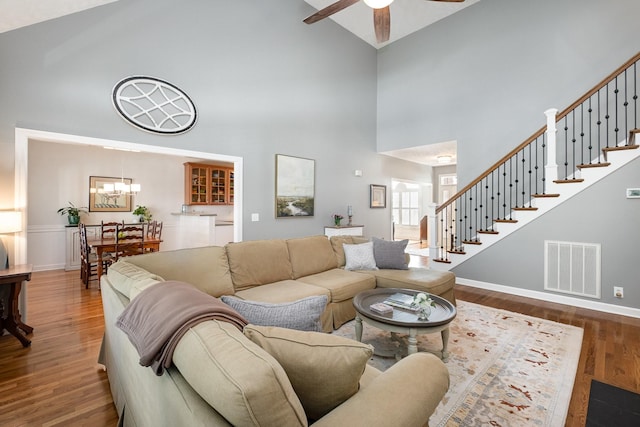 living area with visible vents, ceiling fan with notable chandelier, wood finished floors, stairs, and high vaulted ceiling
