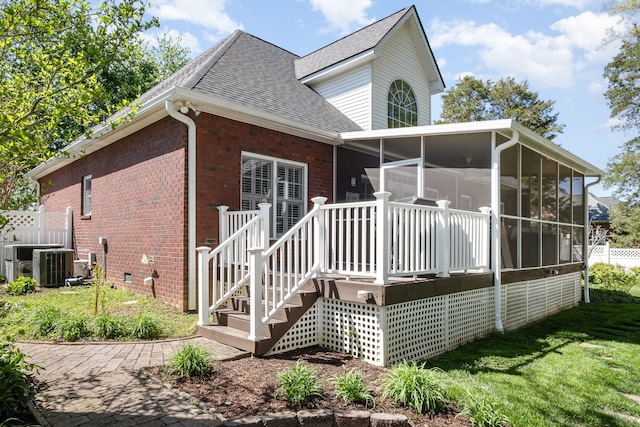 back of property featuring a shingled roof, a sunroom, a lawn, cooling unit, and brick siding