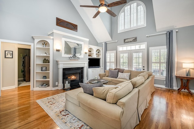 living area with baseboards, built in features, a tiled fireplace, ceiling fan, and high vaulted ceiling