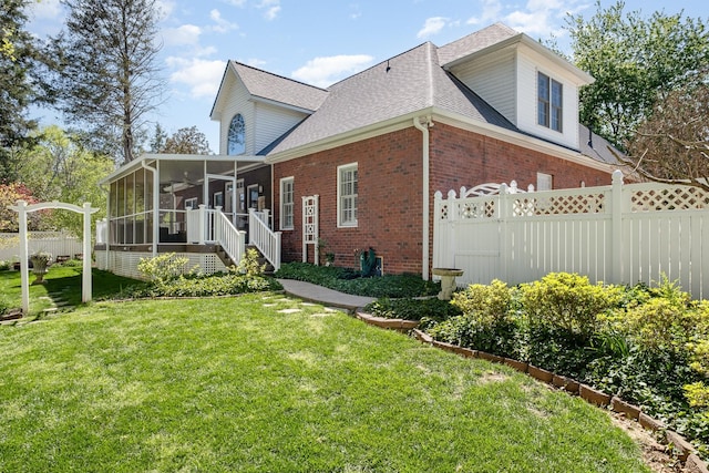 view of side of property with roof with shingles, brick siding, a sunroom, fence, and a lawn