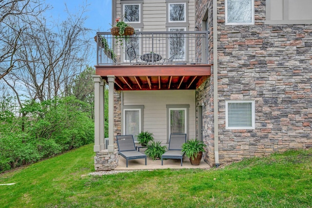 back of property featuring a patio, a balcony, stone siding, and a lawn