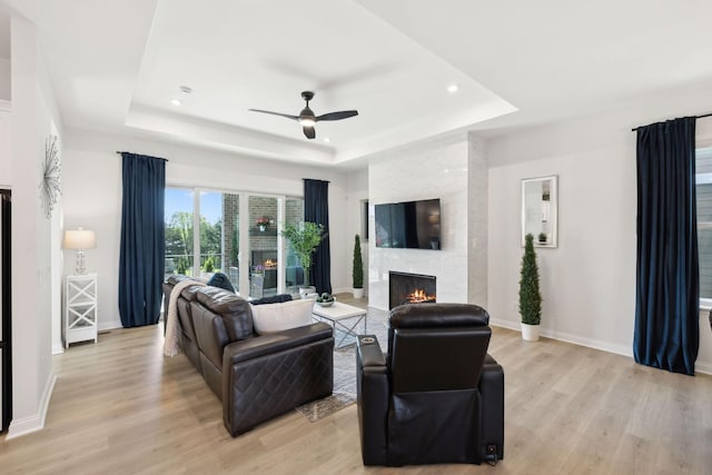 living room featuring a ceiling fan, a large fireplace, light wood-type flooring, baseboards, and a raised ceiling