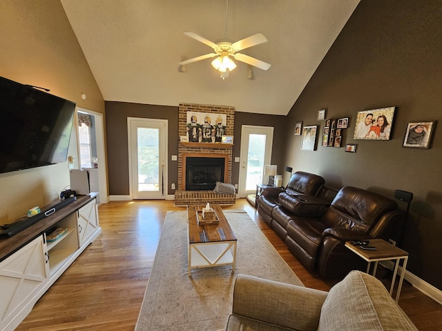 living room featuring a brick fireplace, baseboards, light wood-type flooring, a ceiling fan, and high vaulted ceiling