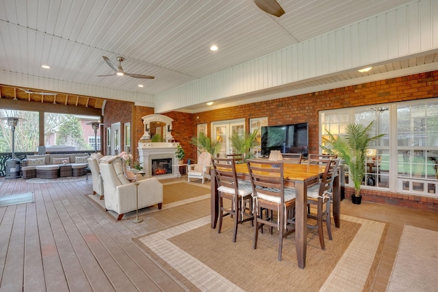 dining area featuring a warm lit fireplace, light wood-type flooring, a ceiling fan, and brick wall