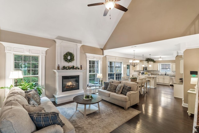 living area featuring ceiling fan with notable chandelier, high vaulted ceiling, dark wood finished floors, and a glass covered fireplace