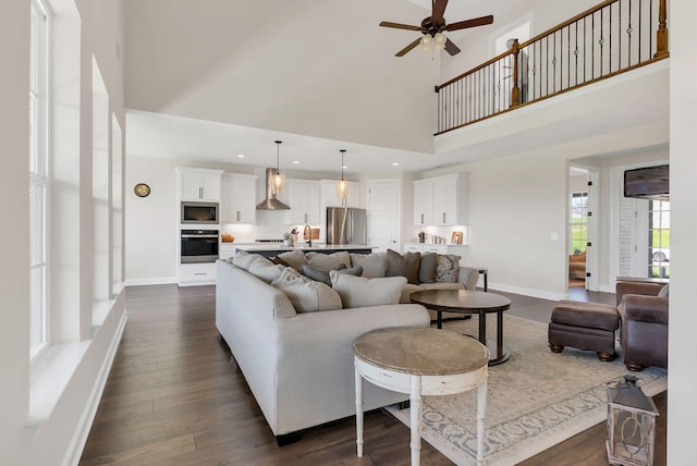 living room featuring dark wood-type flooring, baseboards, a ceiling fan, and a towering ceiling