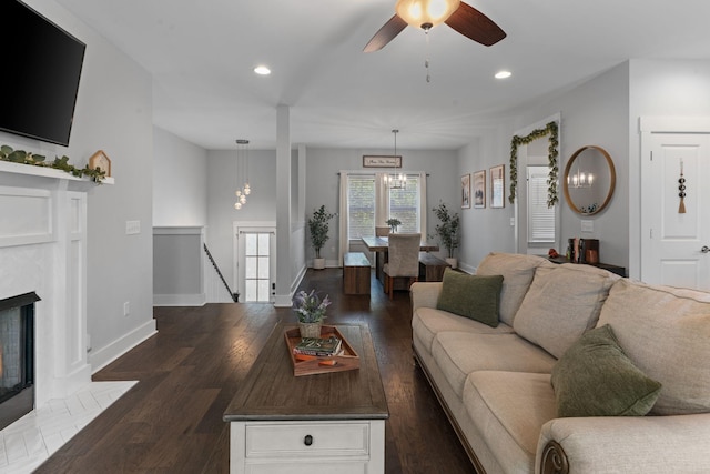 living room featuring dark wood-style flooring, baseboards, recessed lighting, a fireplace with flush hearth, and ceiling fan with notable chandelier
