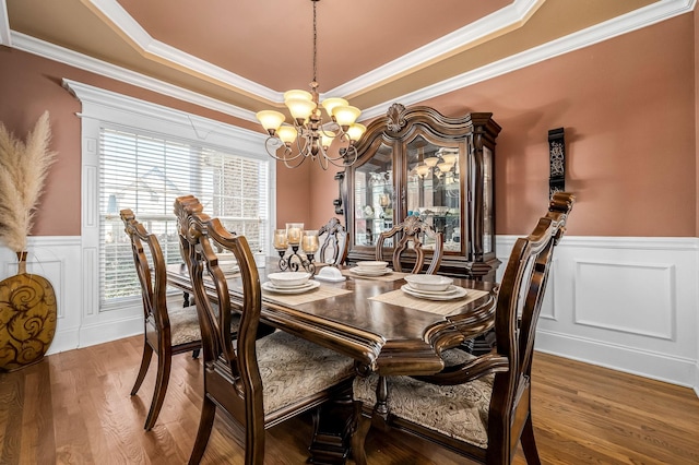 dining area with a raised ceiling, ornamental molding, a chandelier, wainscoting, and wood finished floors