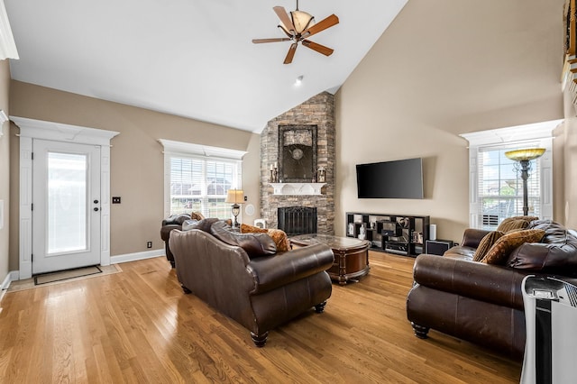 living area featuring high vaulted ceiling, light wood-style flooring, a ceiling fan, a stone fireplace, and baseboards
