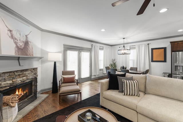 living room with crown molding, wood finished floors, ceiling fan with notable chandelier, and a stone fireplace