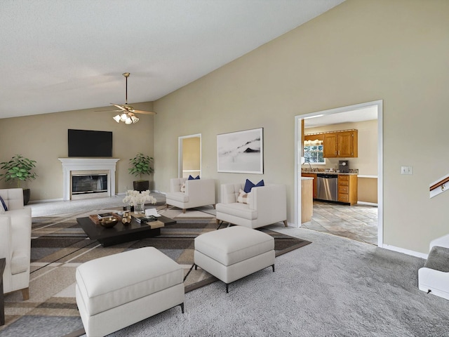 living area featuring baseboards, ceiling fan, high vaulted ceiling, a glass covered fireplace, and light carpet