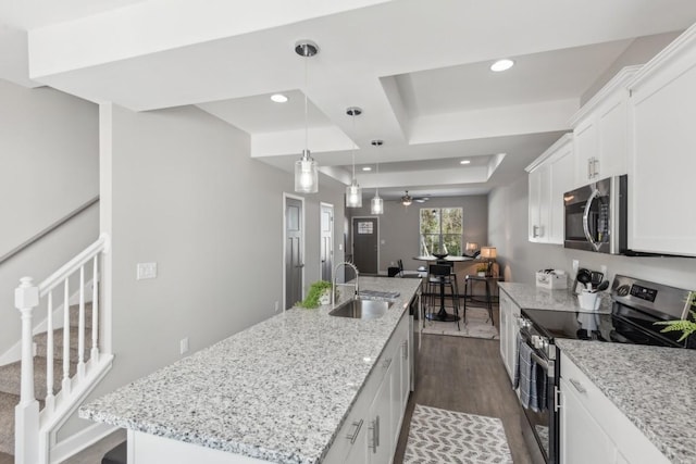 kitchen with a raised ceiling, a sink, appliances with stainless steel finishes, a kitchen island with sink, and dark wood finished floors
