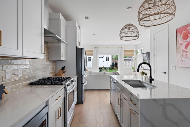 kitchen featuring wall chimney exhaust hood, a sink, open floor plan, appliances with stainless steel finishes, and light wood-style floors