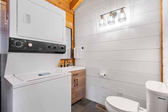 laundry area with visible vents, dark tile patterned floors, stacked washer and clothes dryer, laundry area, and wood walls