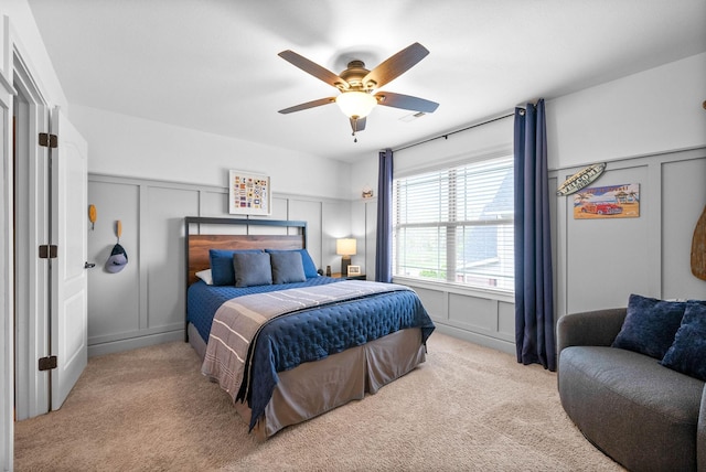 carpeted bedroom with a decorative wall, wainscoting, and ceiling fan