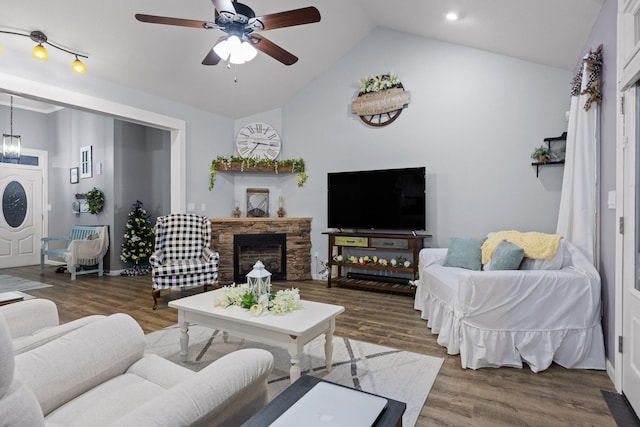 living room featuring ceiling fan with notable chandelier, wood finished floors, a stone fireplace, and vaulted ceiling