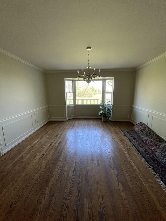 unfurnished dining area featuring dark wood-style flooring, ornamental molding, a chandelier, a wainscoted wall, and a decorative wall