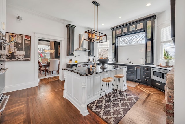 kitchen featuring wall chimney range hood, a sink, glass insert cabinets, stainless steel microwave, and a notable chandelier
