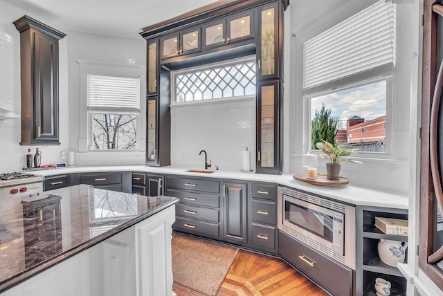 kitchen featuring a sink, stainless steel microwave, light wood-style floors, glass insert cabinets, and light stone countertops
