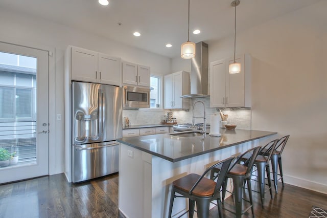 kitchen featuring stainless steel appliances, wall chimney exhaust hood, dark countertops, a peninsula, and a kitchen bar