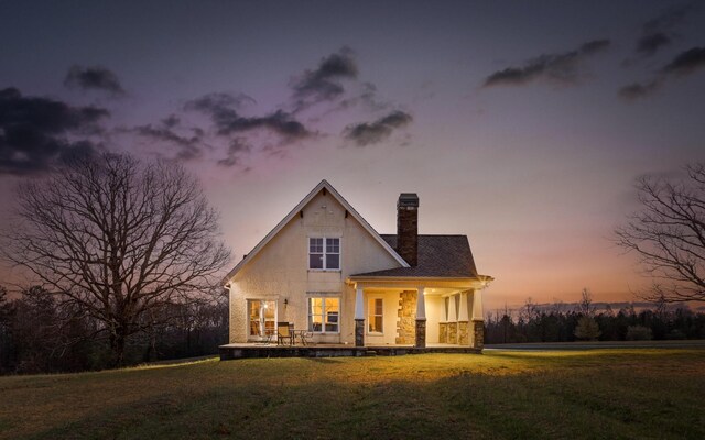 back of house with stucco siding, stone siding, a chimney, and a lawn