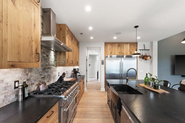 kitchen with light wood-style flooring, dark countertops, high end appliances, wall chimney exhaust hood, and a sink