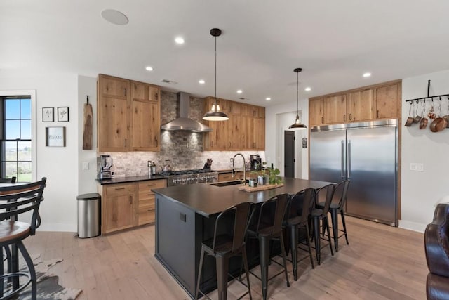 kitchen with wall chimney exhaust hood, a sink, dark countertops, built in fridge, and light wood-style flooring